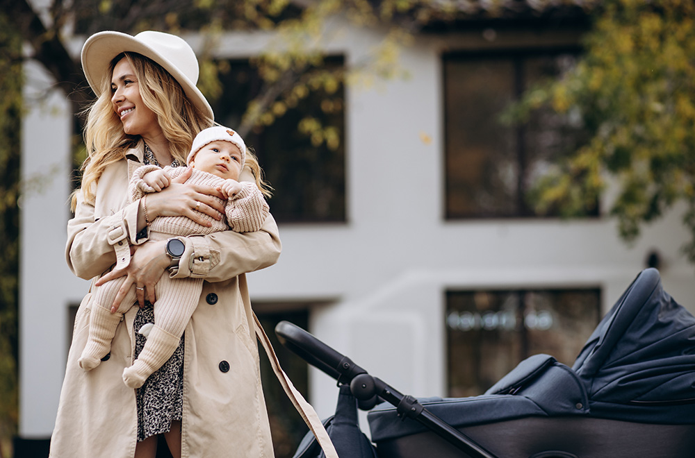 Naine hoiab süles beebit ja kõnnib vankri kõrval hoone taustal